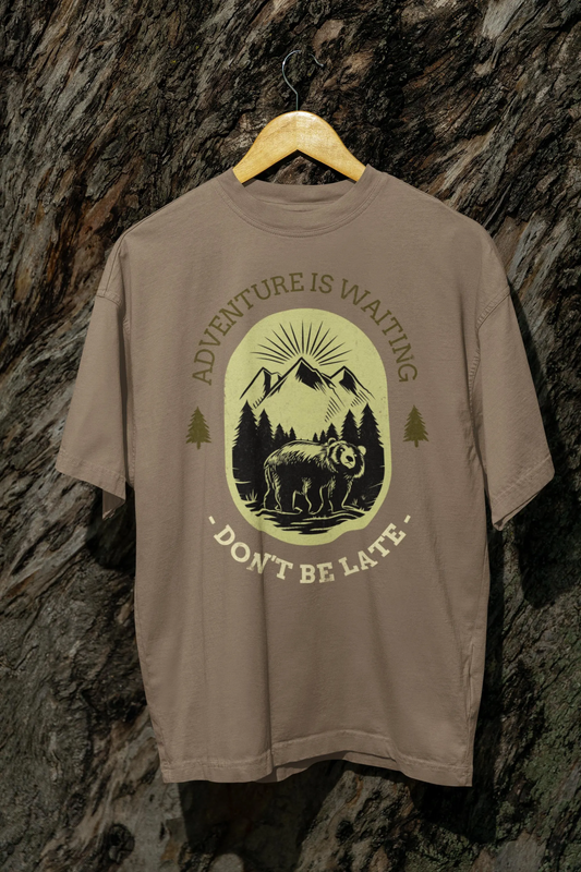 Beige t-shirt with bear graphic and text 'Adventure is waiting, Don't be late' on a stone wall background