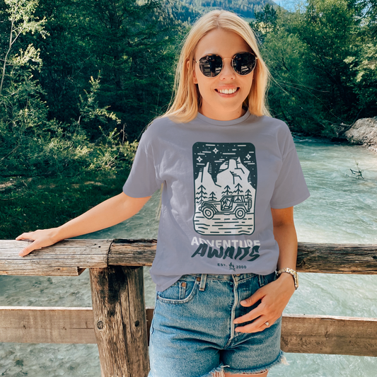 Woman wearing a gray t-shirt with a graphic design, standing by a wooden railing with a scenic background.
