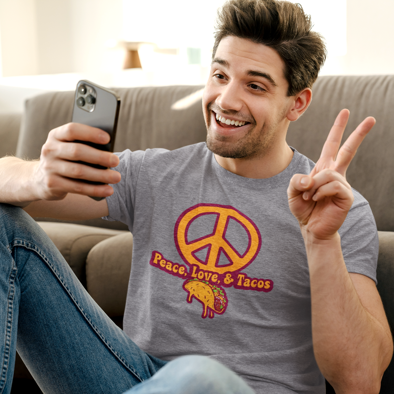 Man sitting on a couch wearing a 'Peace Love & Tacos' shirt, holding a phone and making a peace sign.