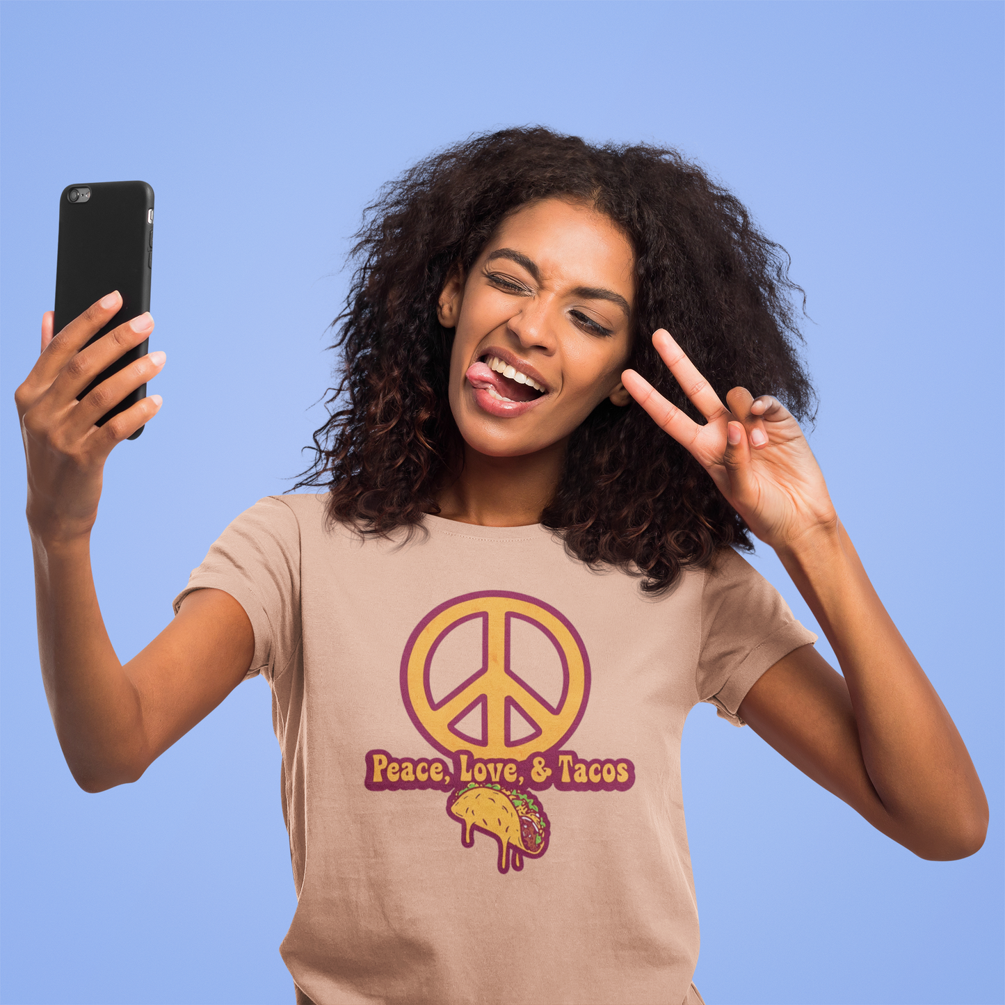 Woman wearing a 'Peace, Love, & Tacos' t-shirt, taking a selfie against a blue background