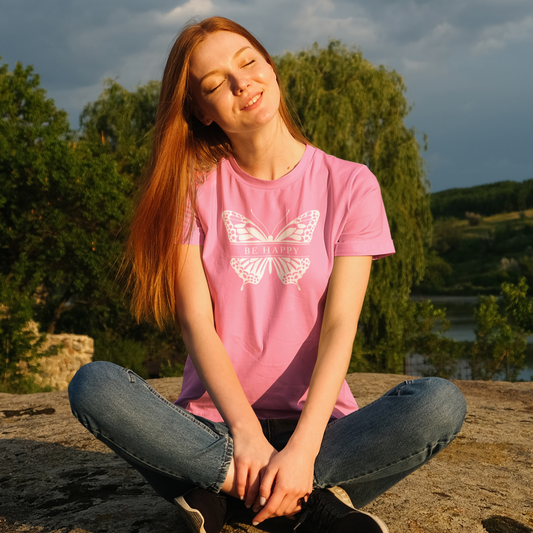 Person sitting outdoors wearing a pink t-shirt with a butterfly design.
