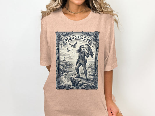Graphic tee with woman holding a bird, coastal scene, and "Weird Girls Club" text.