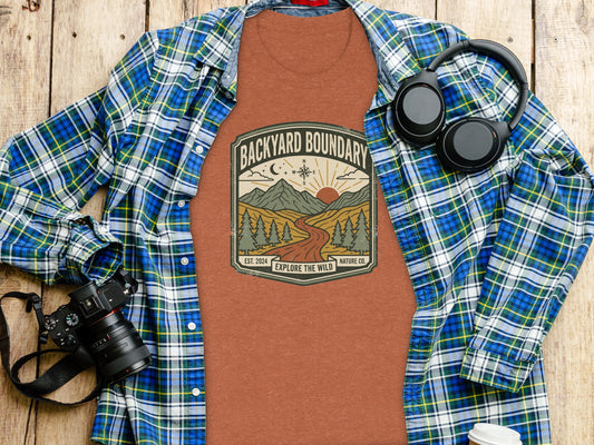 Rugged Backyard Boundary: Explore The Wild Wilderness Shirt: Brown t-shirt with "Backyard Boundary" mountain landscape graphic, headphones, and camera.