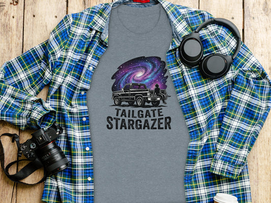 Heritage Cosmic Tailgate Stargazer Nomad Tee: T-shirt with "Stargate Stargazer" text and galaxy graphic, laid flat with a camera and headphones.