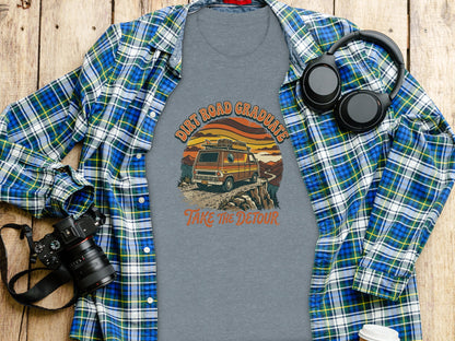 Heritage Dirt Road Graduate Adventure Tee Nomad Tee: Flannel shirt, t-shirt with "Dirt Road Graduate" van graphic, headphones, and camera on wood.