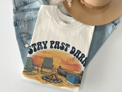 Cream t-shirt with "Stay Past Dark" graphic featuring campfire, chairs, and sunset.
