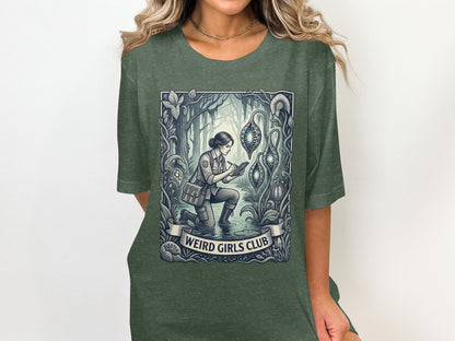 T-shirt graphic of a woman in a forest, examining glowing pods, with "Weird Girls Club" text.