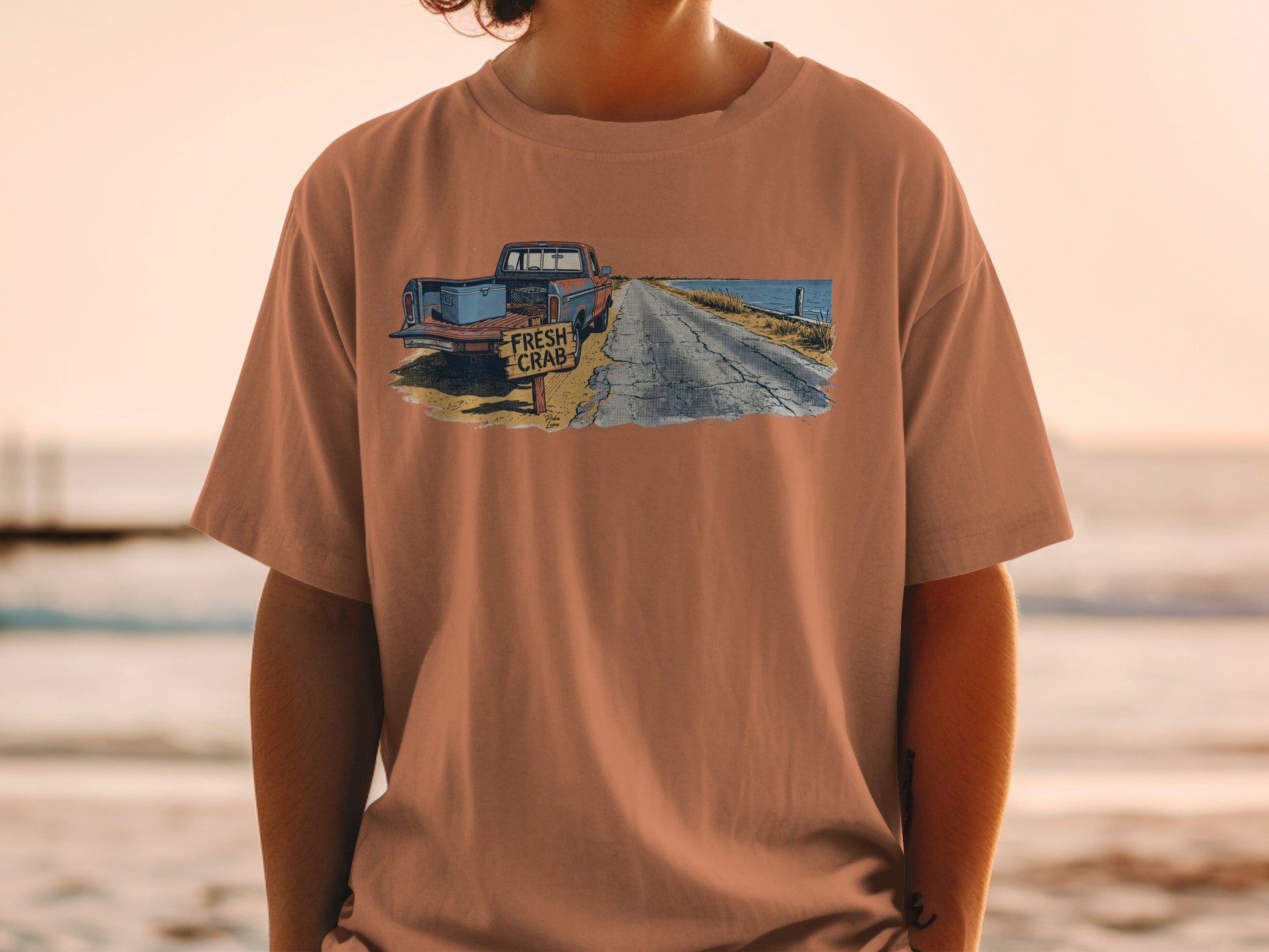 T-shirt graphic of a rusty truck, crab sign, and coastal road at sunset.