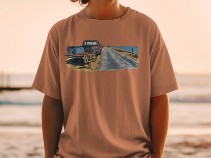 T-shirt graphic of a rusty truck, crab sign, and coastal road at sunset.
