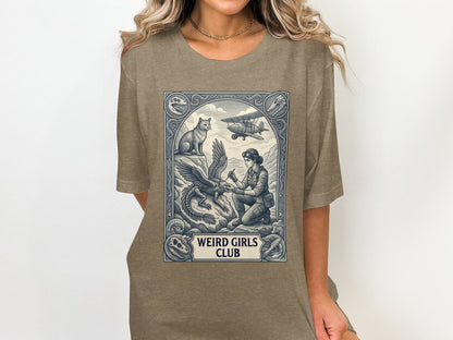 T-shirt graphic with woman, dragon, cat, and biplane in ornate frame with "Weird Girls Club" text.