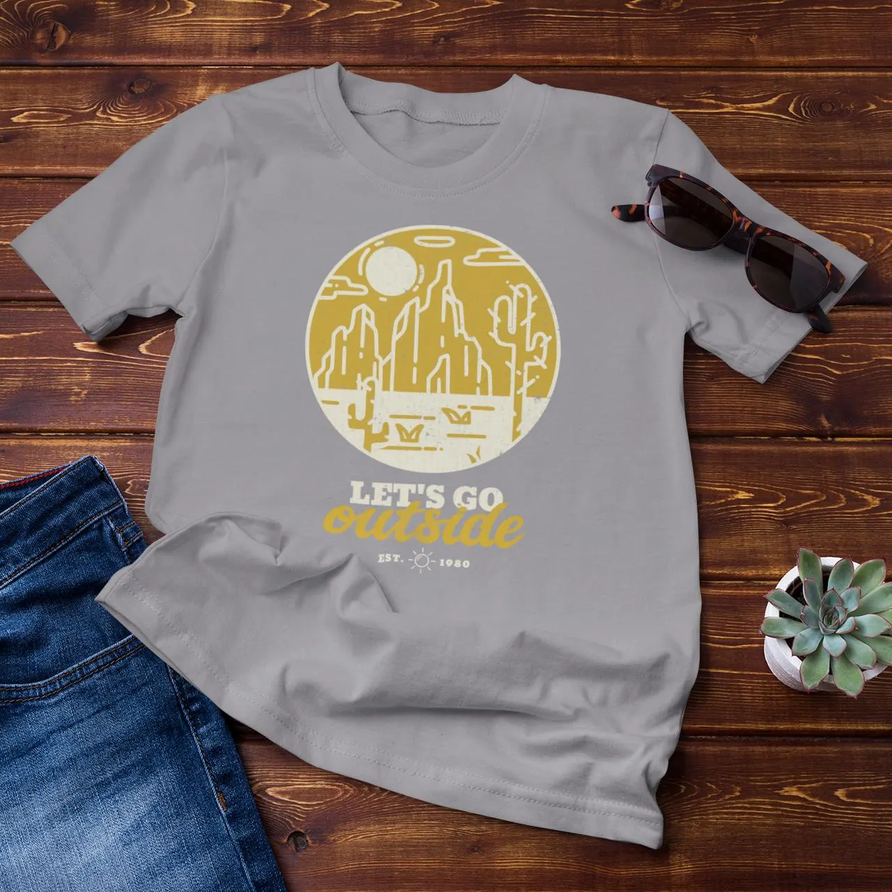 Gray t-shirt with a yellow graphic and text on a wooden surface with jeans and sunglasses.