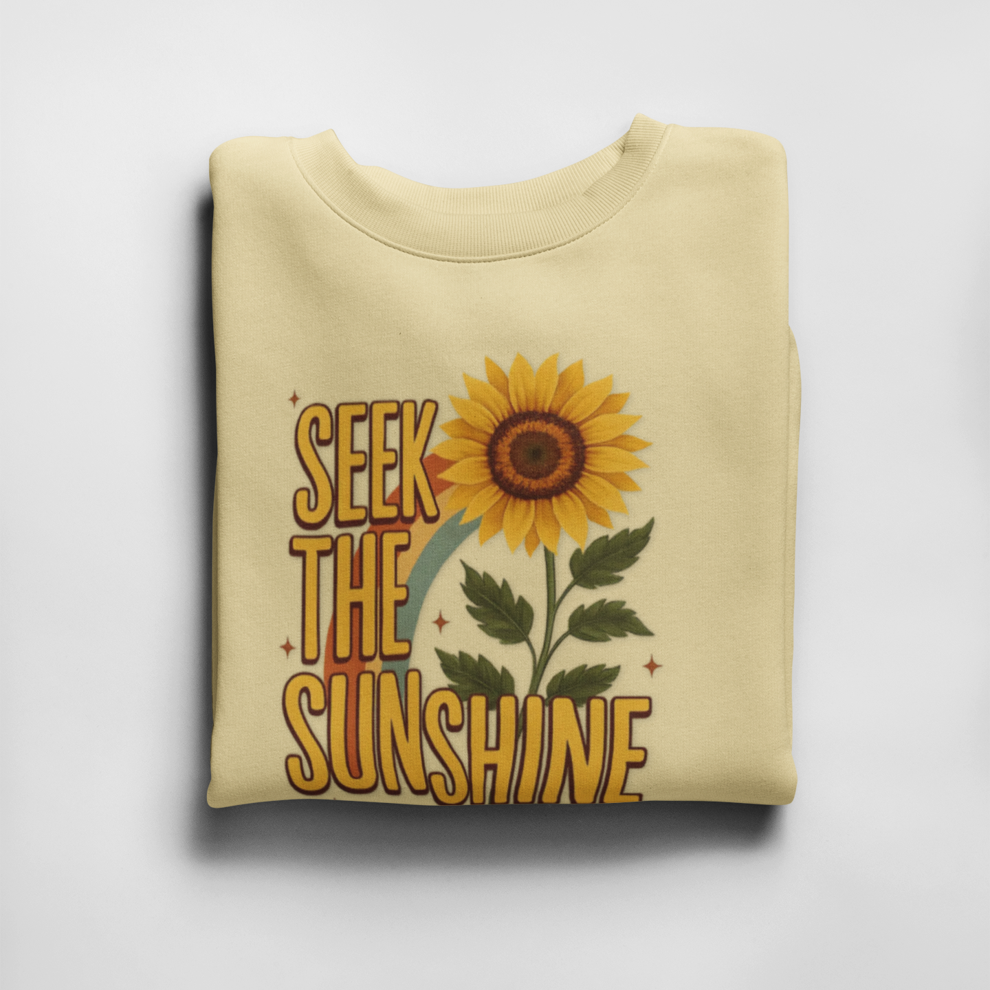 Folded yellow sweatshirt with sunflower graphic and 'Seek the Sunshine' text on a light gray background