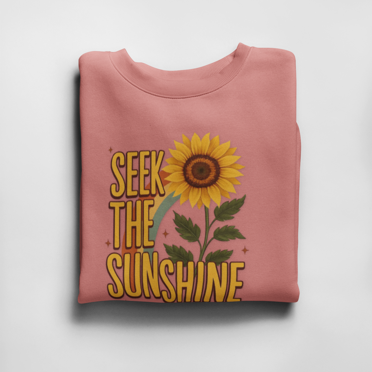 Folded pink sweatshirt with sunflower and 'Seek the Sunshine' text on a light gray background