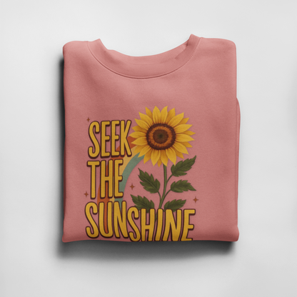 Folded pink sweatshirt with sunflower and 'Seek the Sunshine' text on a light gray background