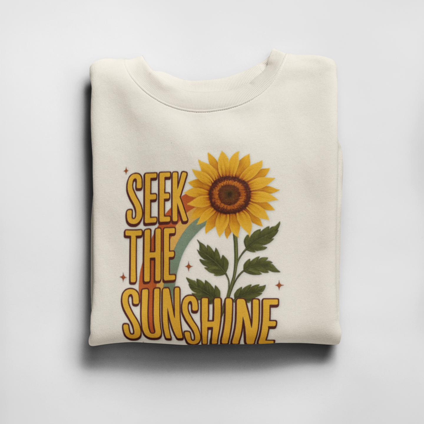 Folded white sweatshirt with sunflower and 'Seek the Sunshine' text on a light gray background