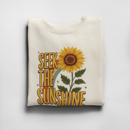 Folded white sweatshirt with sunflower and 'Seek the Sunshine' text on a light gray background