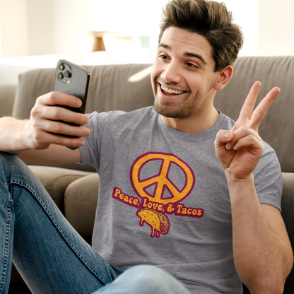 Man sitting on a couch wearing a 'Peace Love & Tacos' shirt, holding a phone and making a peace sign.