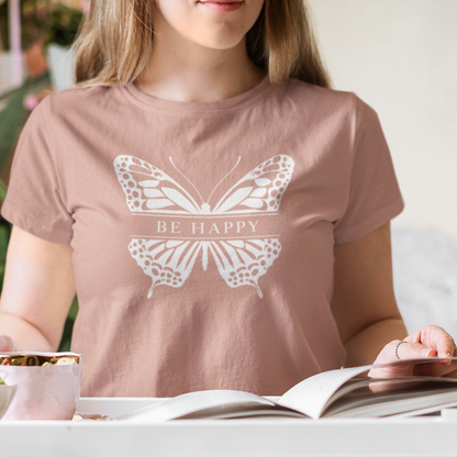 Person wearing a t-shirt with a butterfly design and 'Be Happy' text, sitting at a table with a bowl of fruit and an open book.