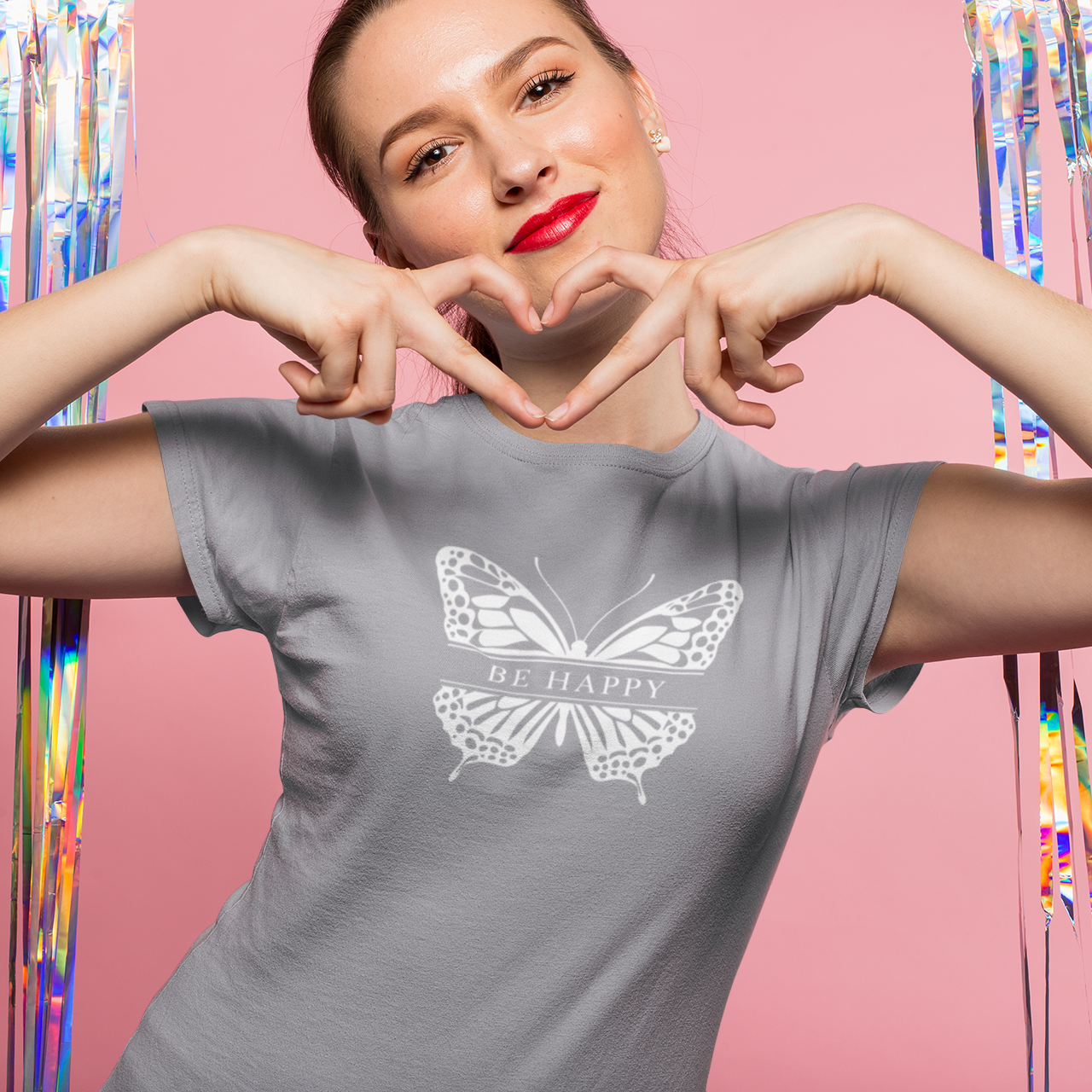 Woman wearing a gray t-shirt with a butterfly design and 'Be Happy' text, making a heart shape with her hands against a colorful curtain background.