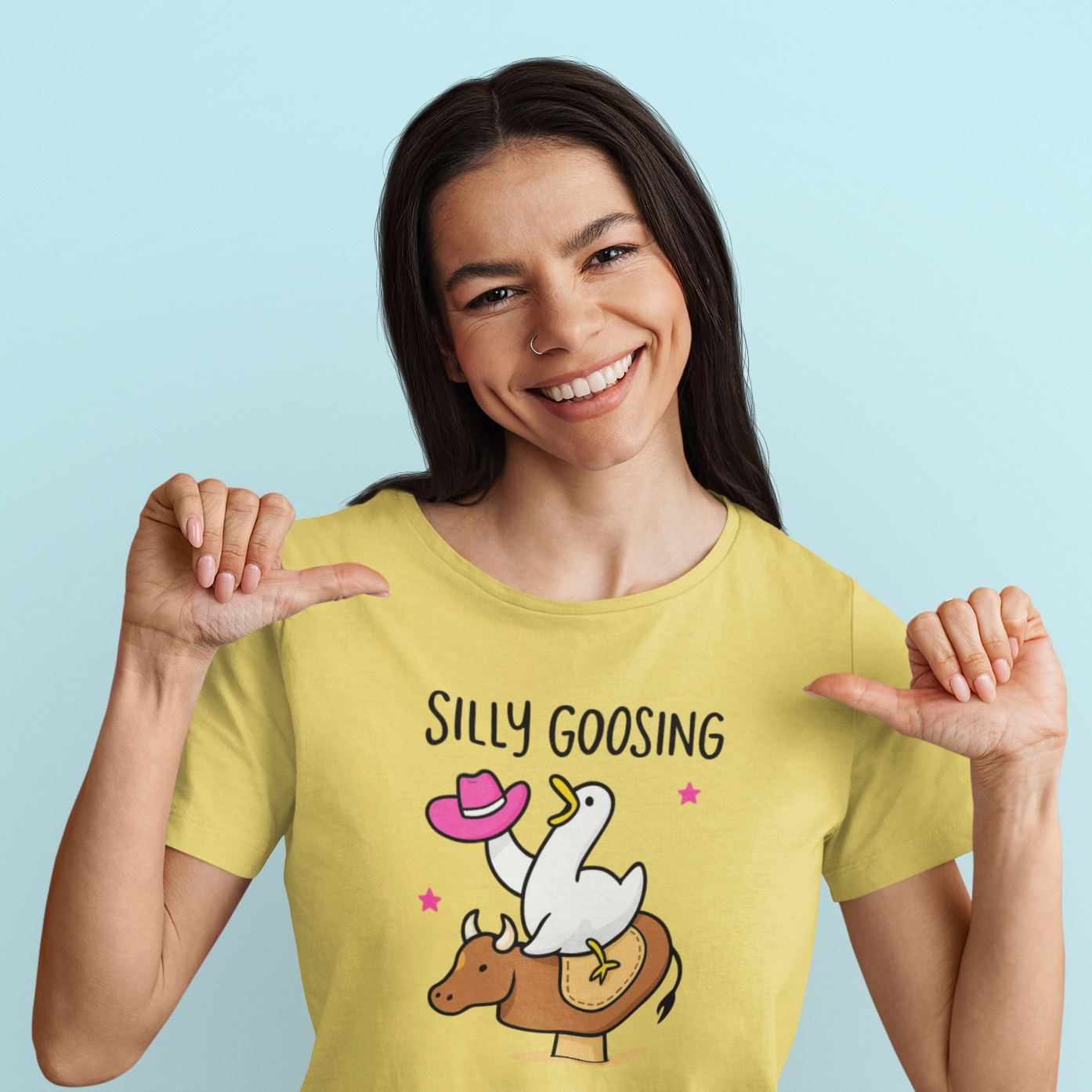 Woman wearing a yellow t-shirt with a graphic design and text, smiling and pointing at the shirt against a light blue background.
