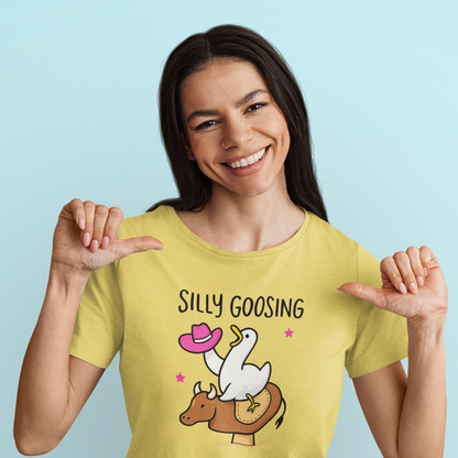 Woman wearing a yellow t-shirt with a graphic design and text, smiling and pointing at the shirt against a light blue background.