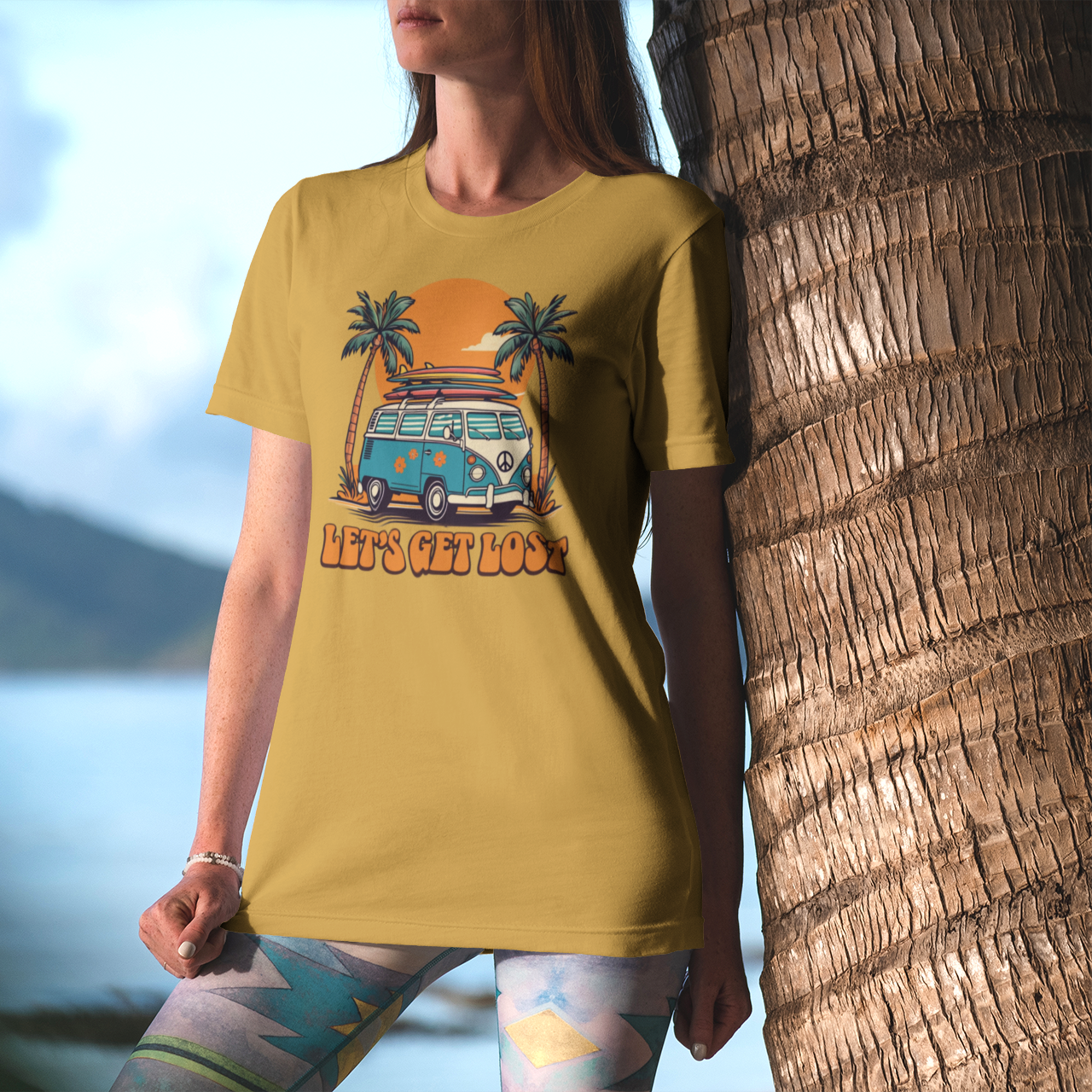 Person wearing a yellow t-shirt with a graphic design of a van and palm trees, standing next to a palm tree with a mountain and lake in the background.