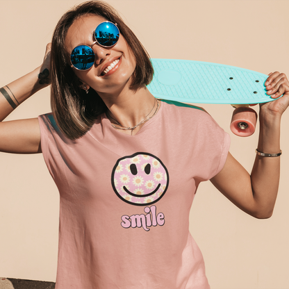 Person wearing a pink t-shirt with a smiley face design, holding a teal skateboard against a beige background.