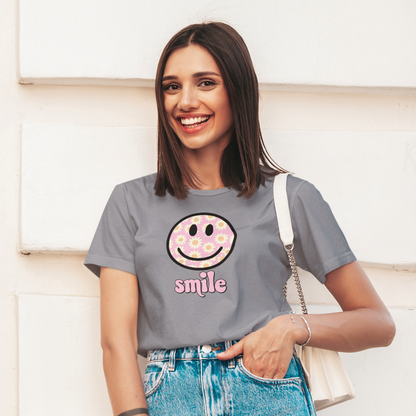 Woman wearing a gray t-shirt with a smiley face design and denim shorts, standing against a light-colored wall.