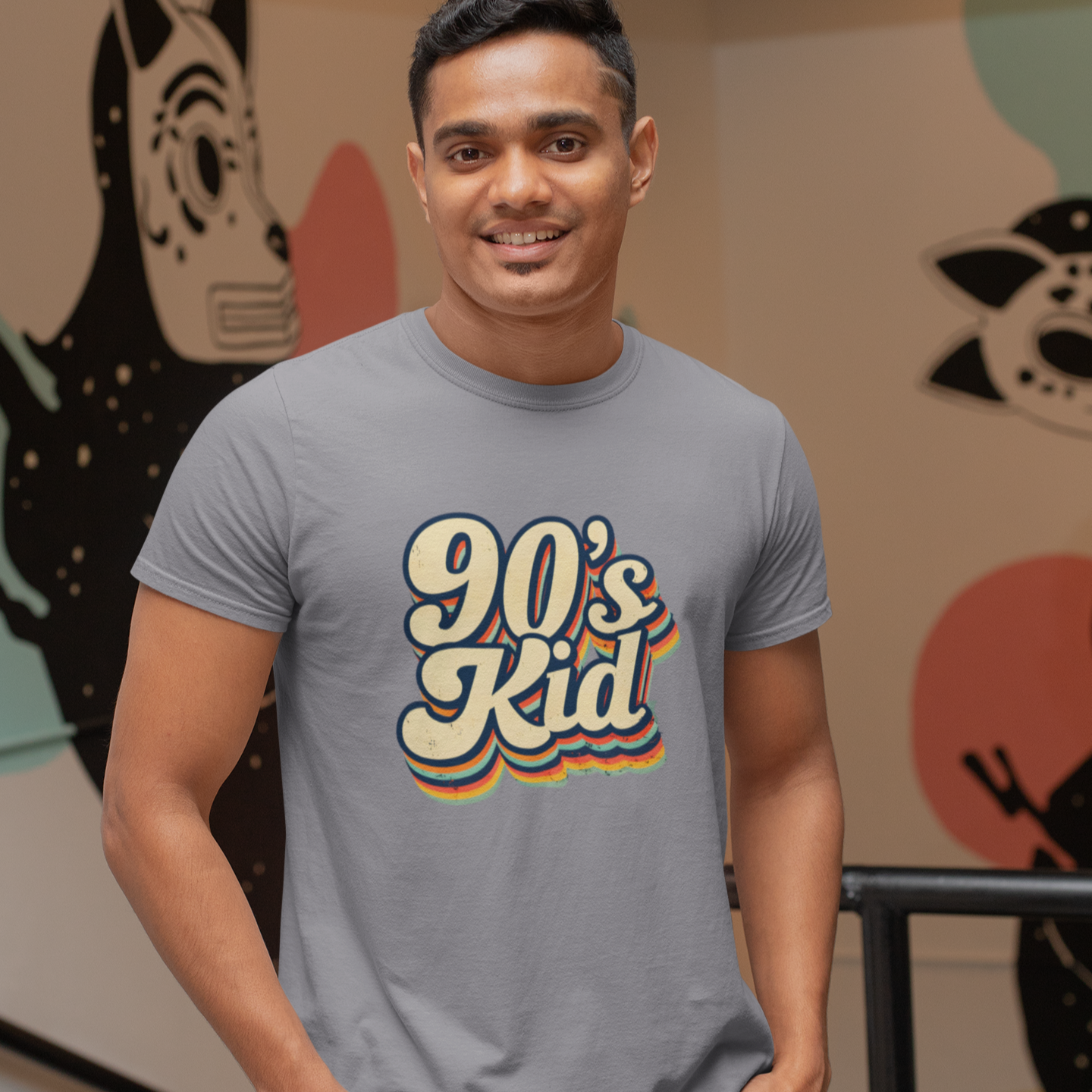 Man wearing a gray t-shirt with '90's Kid' design in front of colorful abstract wall art.