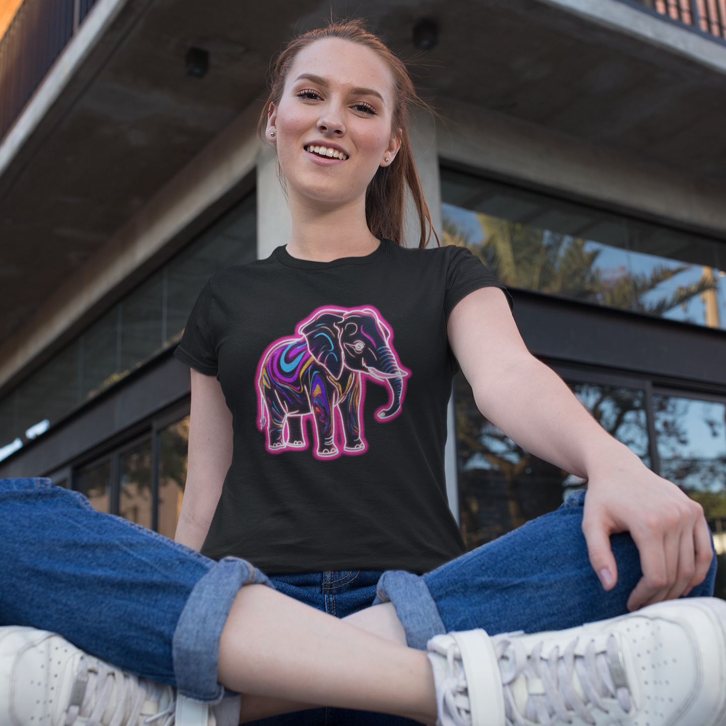 Person wearing a black t-shirt with an elephant design, sitting on a bench in front of a building.