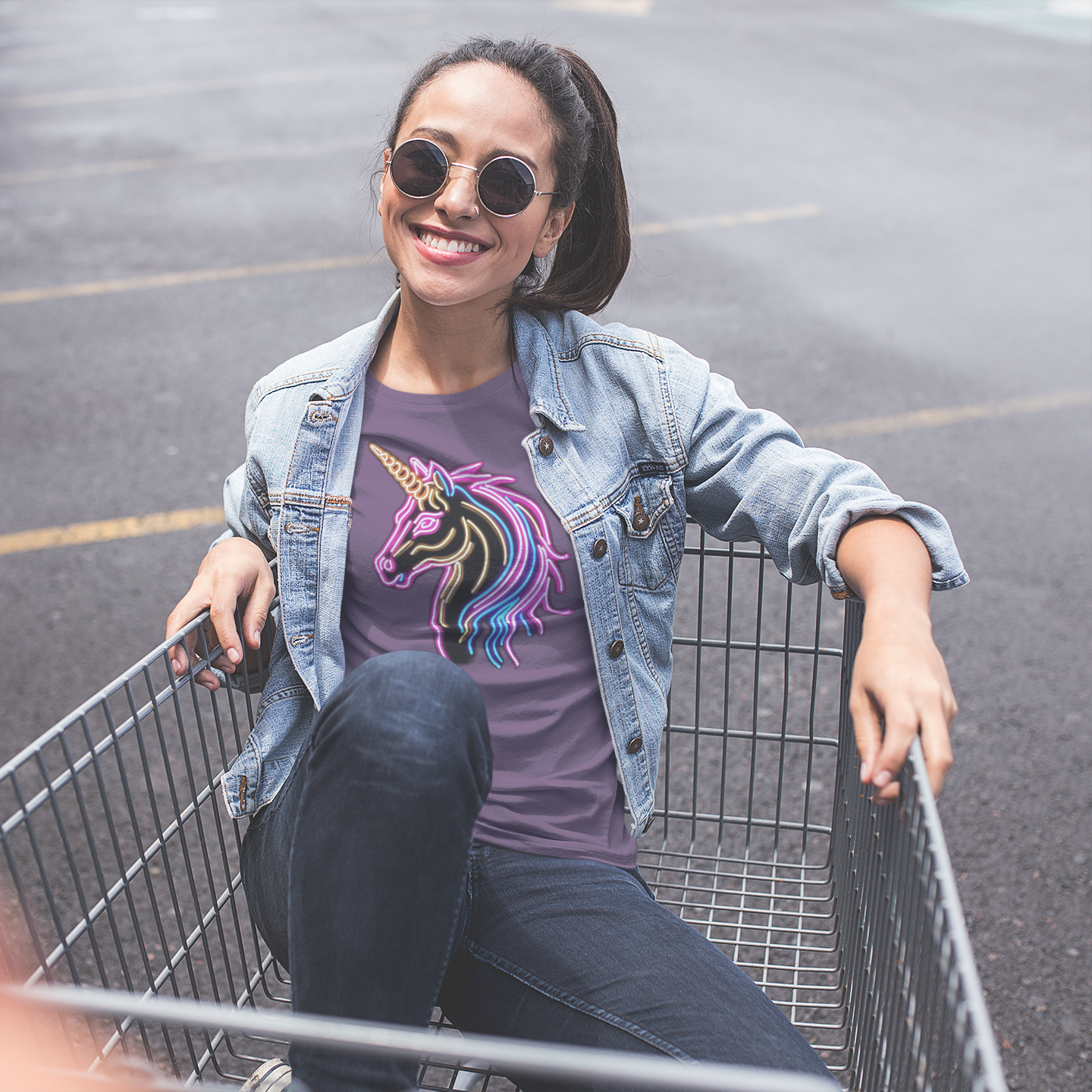 Woman sitting in a shopping cart wearing a t-shirt with a unicorn design in an outdoor setting.