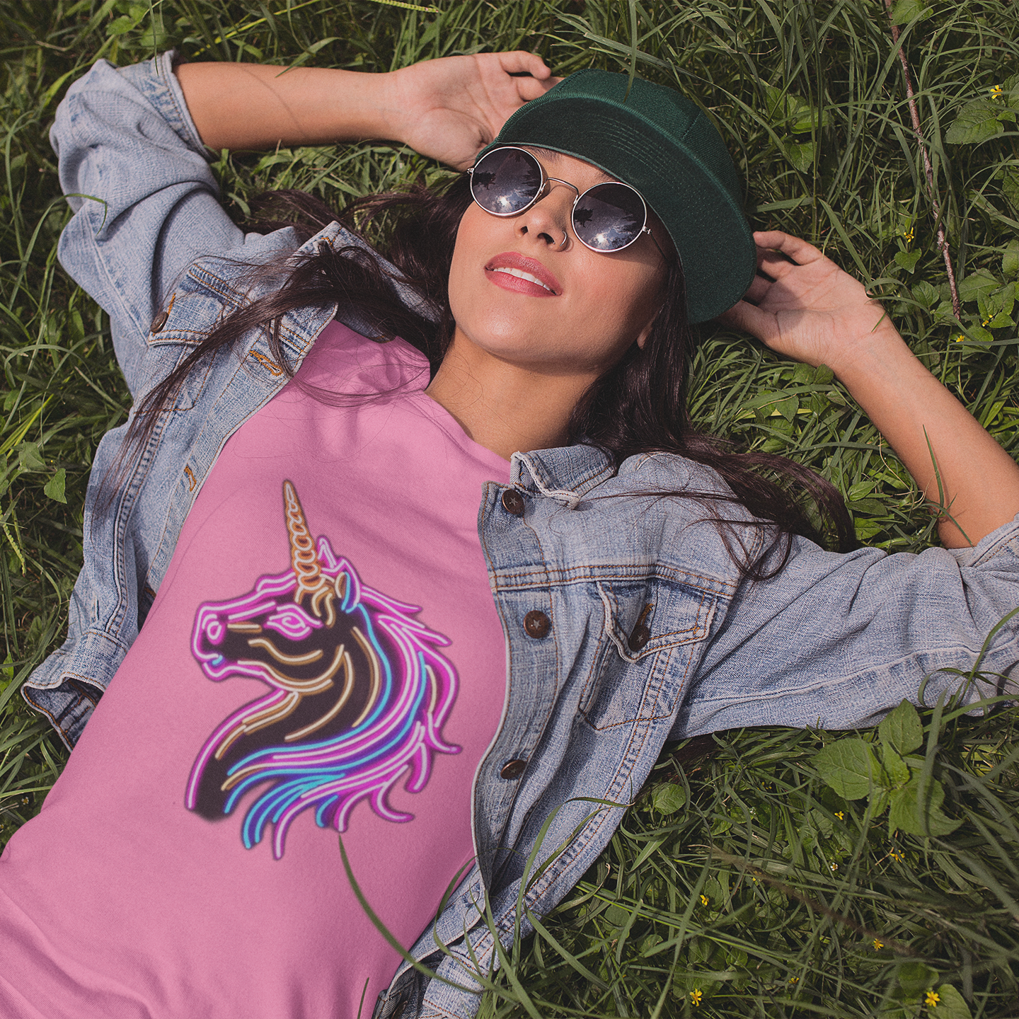 Woman wearing a pink shirt with a unicorn design, denim jacket, and green hat lying on grass.