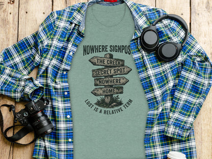 Nowhere Signpost Adventure: Backcountry Outpost Shirt: Green t-shirt with "Nowhere Signpost" graphic and directional arrows, laid flat with headphones and camera.
