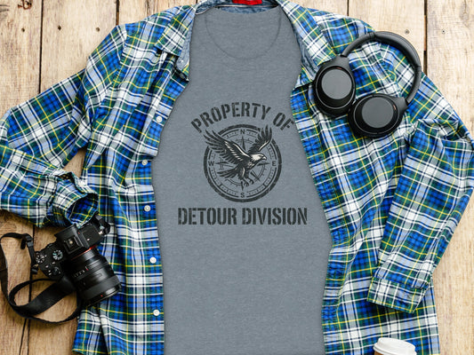 Rugged Detour Division Compass Eagle Tee Wilderness Shirt: Grey t-shirt with "PROPERTY OF DETOUR DIVISION" and compass design, on wooden surface with headphones.