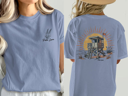 Periwinkle t-shirt with Puka Luna logo and a lifeguard stand at sunset graphic.