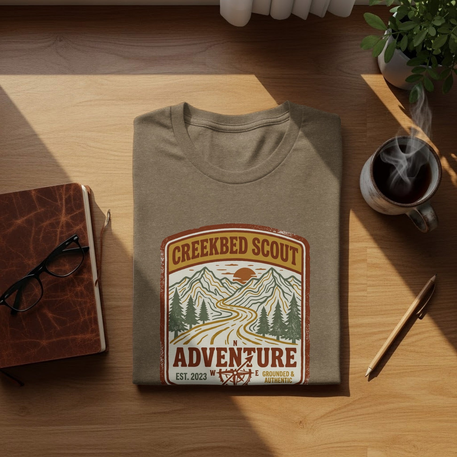 Rugged Creekbed Scout Adventure Emblem Wilderness Shirt: Brown t-shirt with "Creekbed Scout Adventure" logo featuring mountains and a winding road.