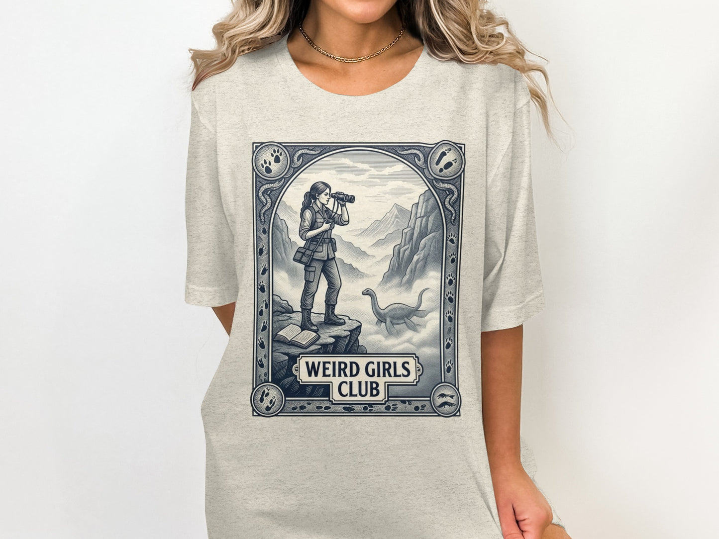 Graphic t-shirt with a woman looking through binoculars near a dinosaur, "Weird Girls Club" text.