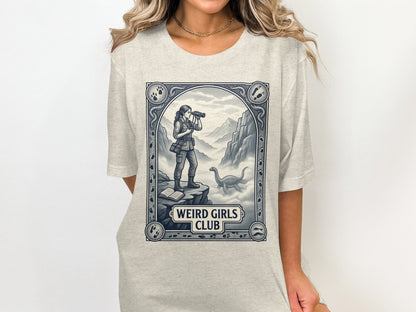 Graphic t-shirt with a woman looking through binoculars near a dinosaur, "Weird Girls Club" text.