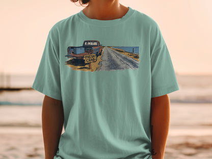T-shirt graphic with rusty truck, crab sign, and ocean road at sunset.