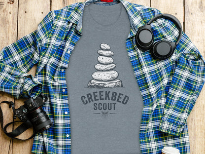 Creekbed Scout Cairn Graphic: The Explorer's Adventure Tee: Grey t-shirt with stacked stones and "CREEKBED SCOUT" text, over a blue and green plaid shirt.