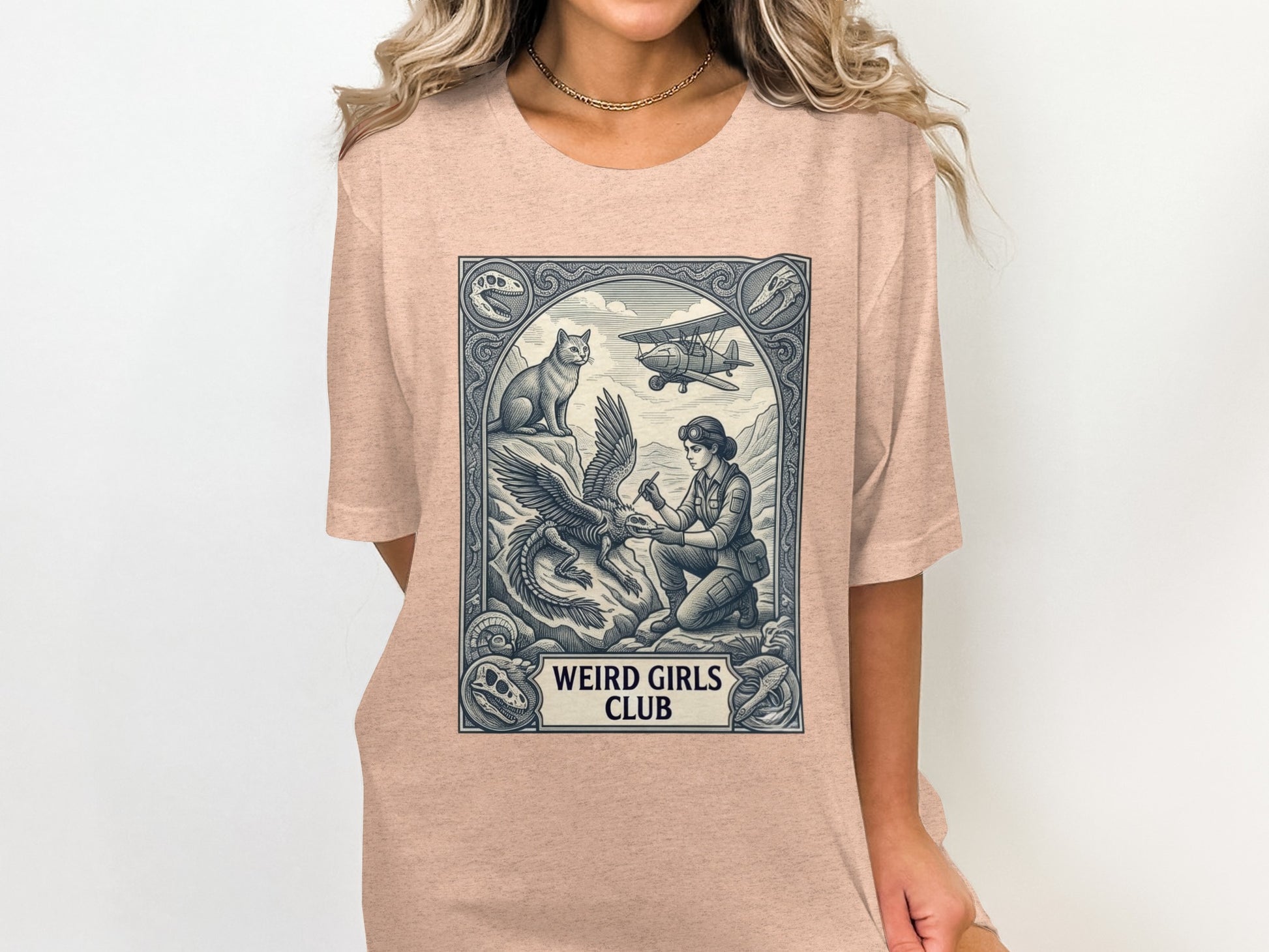 Woman in tan t-shirt with "Weird Girls Club" graphic featuring dragon, cat, and biplane.