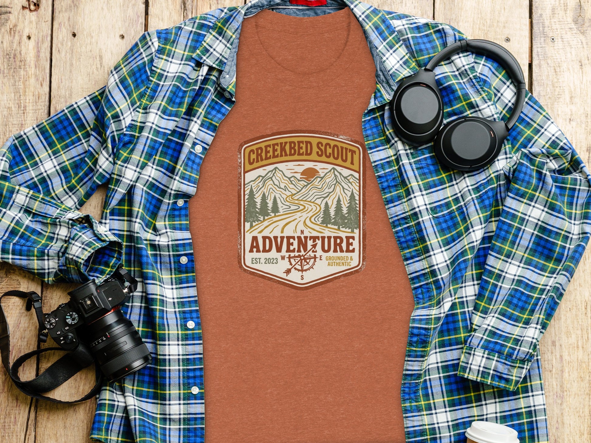 Rugged Creekbed Scout Adventure Emblem Wilderness Shirt: T-shirt with "Creekbed Scout Adventure" logo, laid on a flannel shirt.