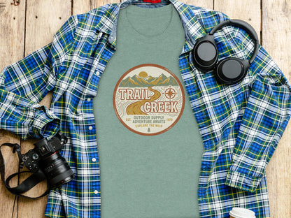Heritage Heritage Explorer Trail Creek Nomad Tee: T-shirt with "Trail Creek Outdoor Supply Adventure Awaits Explore The Wild" circular logo, headphones, and camera.