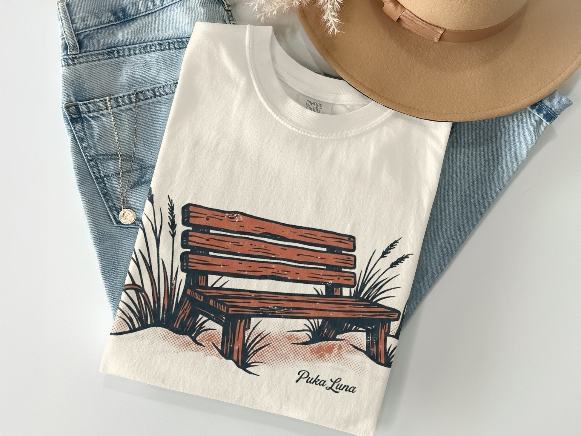 Cream t-shirt with wooden bench and grass graphic, "Puka Luna" text.