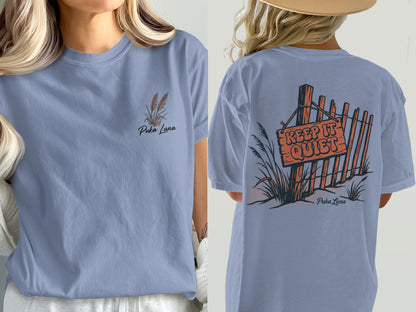 Periwinkle t-shirt with "Puka Luna" text and wheat graphic on front, and fence with "KEEP IT QUIET" sign on back.