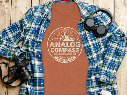 Analog Compass Heritage: The Explorer's Adventure Tee: Orange t-shirt with "Analog Compass Explore the Wild" graphic, laid flat with plaid shirt and headphones.