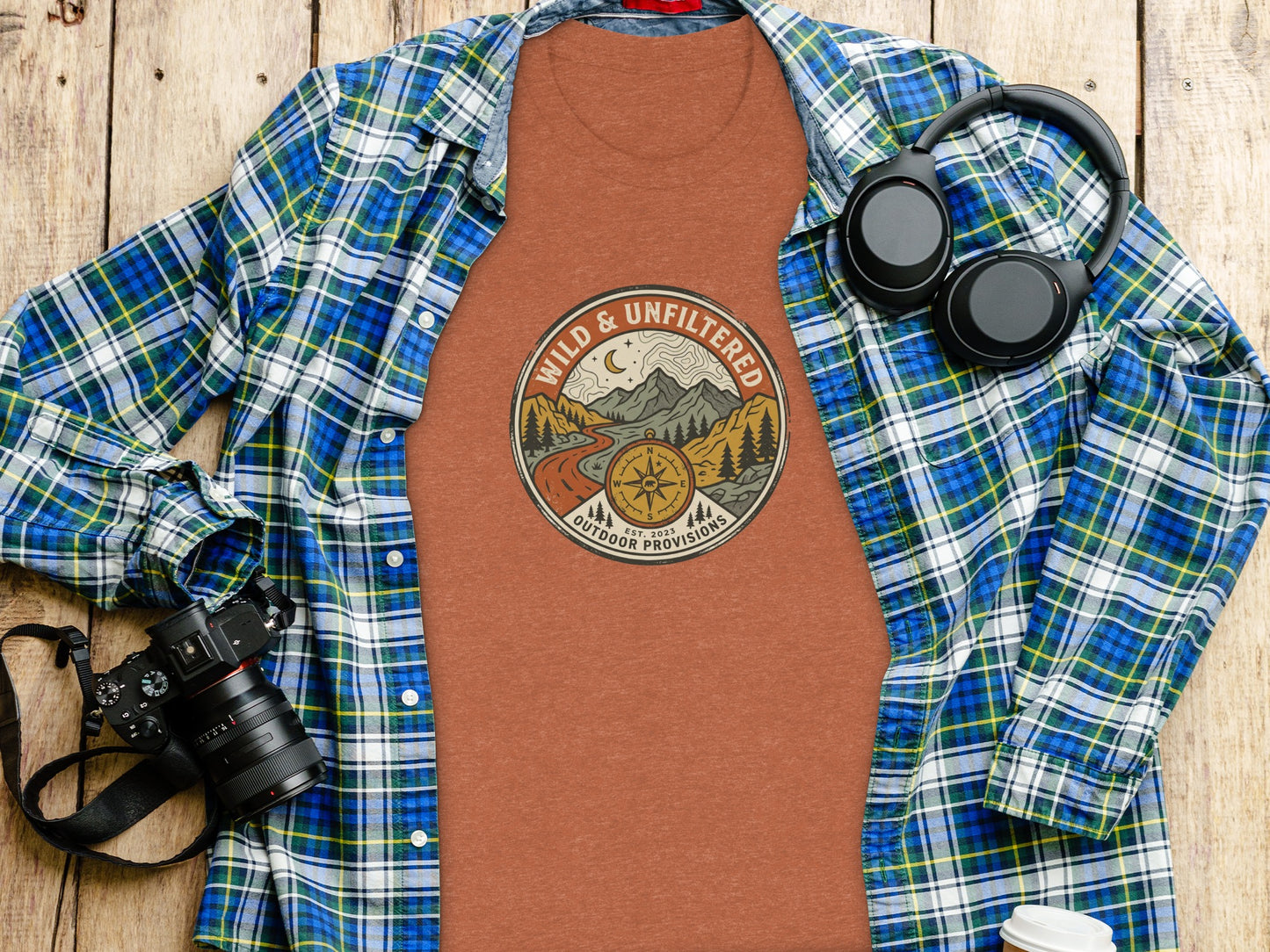 Rugged Wild & Unfiltered Explorer Wilderness Shirt: T-shirt with "Wild & Unfiltered Outdoor Provisions" mountain logo, laid flat with plaid shirt and headphones.