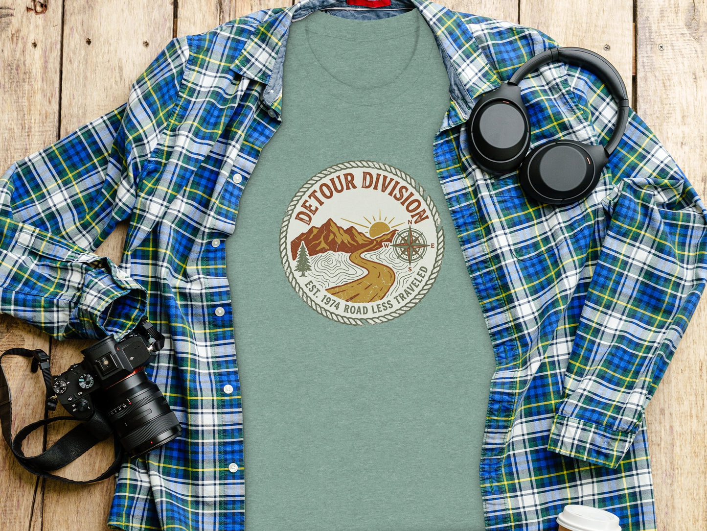 Detour Division Heritage | Vintage Outdoor Apparel: Green t-shirt with "Detour Division" graphic, headphones, and camera on a wooden surface.