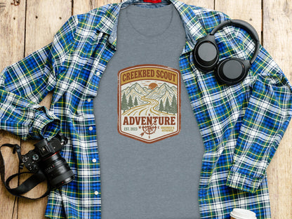 Rugged Creekbed Scout Adventure Emblem Wilderness Shirt: Grey t-shirt with "Creekbed Scout Adventure" logo, laid on a wooden surface.