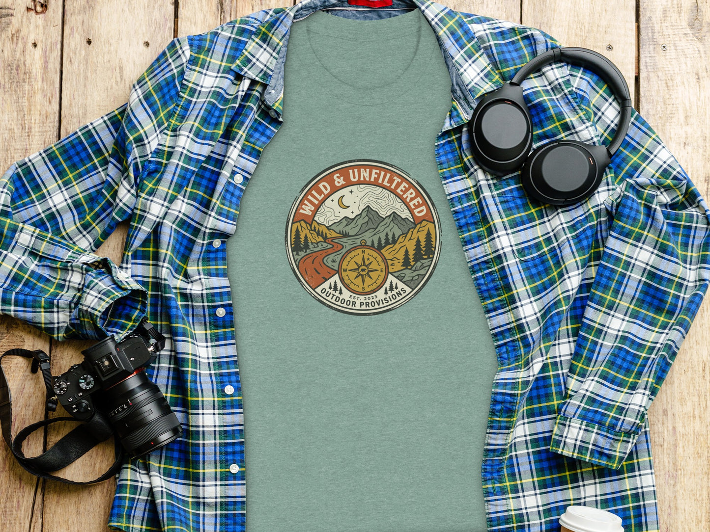 Rugged Wild & Unfiltered Explorer Wilderness Shirt: Green t-shirt with "Wild & Unfiltered Outdoor Provisions" graphic, plaid shirt, headphones, camera.